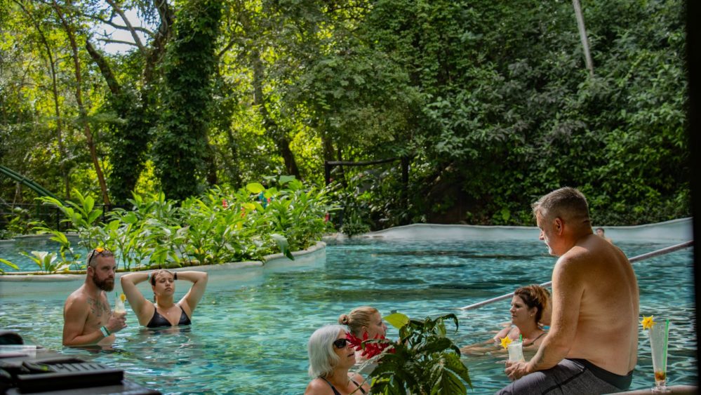 Personas relajándose en una piscina rodeada de bosque, hablando y con bebidas en la mano.
