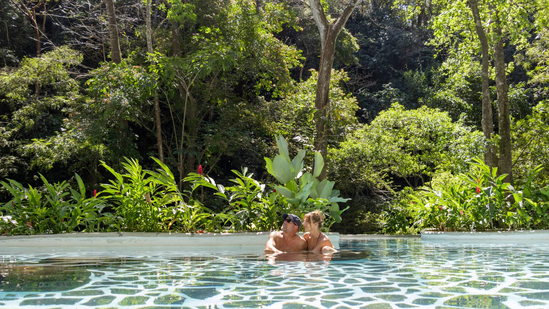 Couple in pool surrounded by lush trees and greenery.