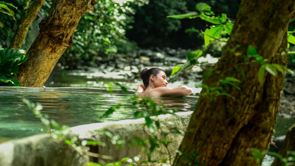 Woman relaxing in a natural pool surrounded by trees and greenery.