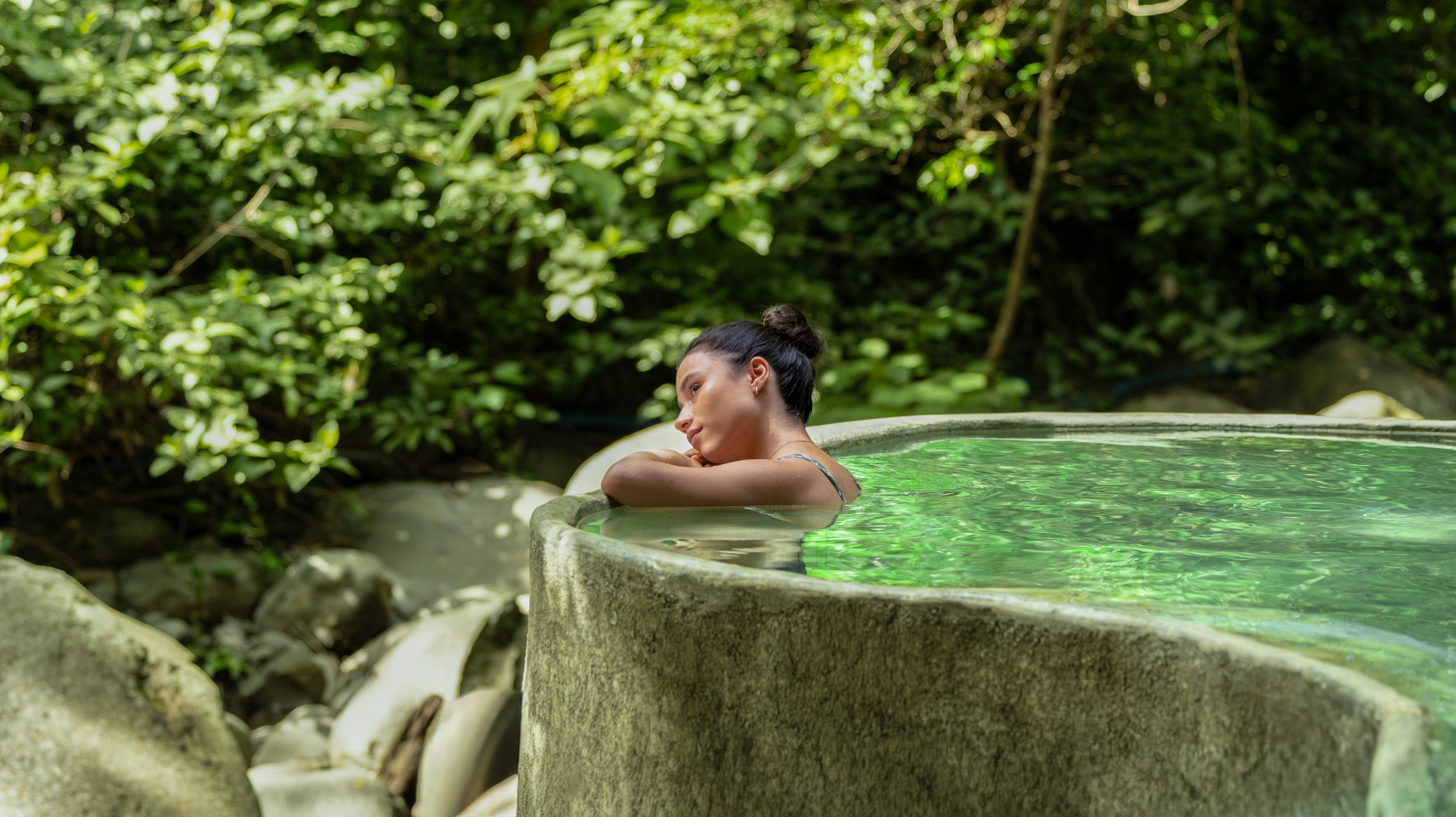 Person relaxing in a stone hot tub surrounded by lush greenery.