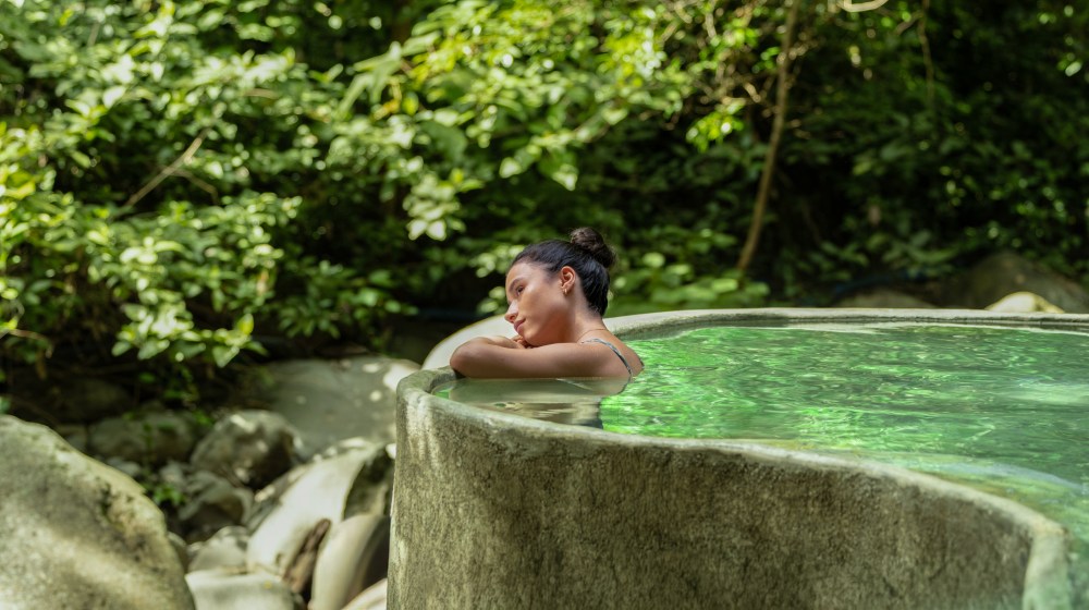 Person relaxing in a natural stone pool surrounded by lush greenery.