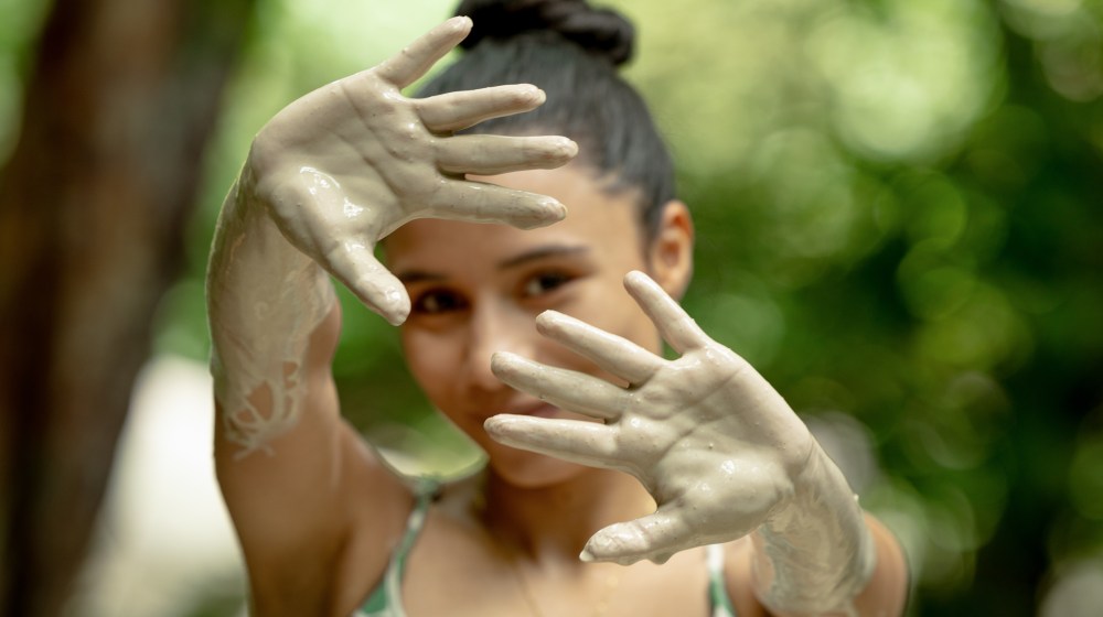 Person with clay-covered hands raised in front of face, green background blurred.