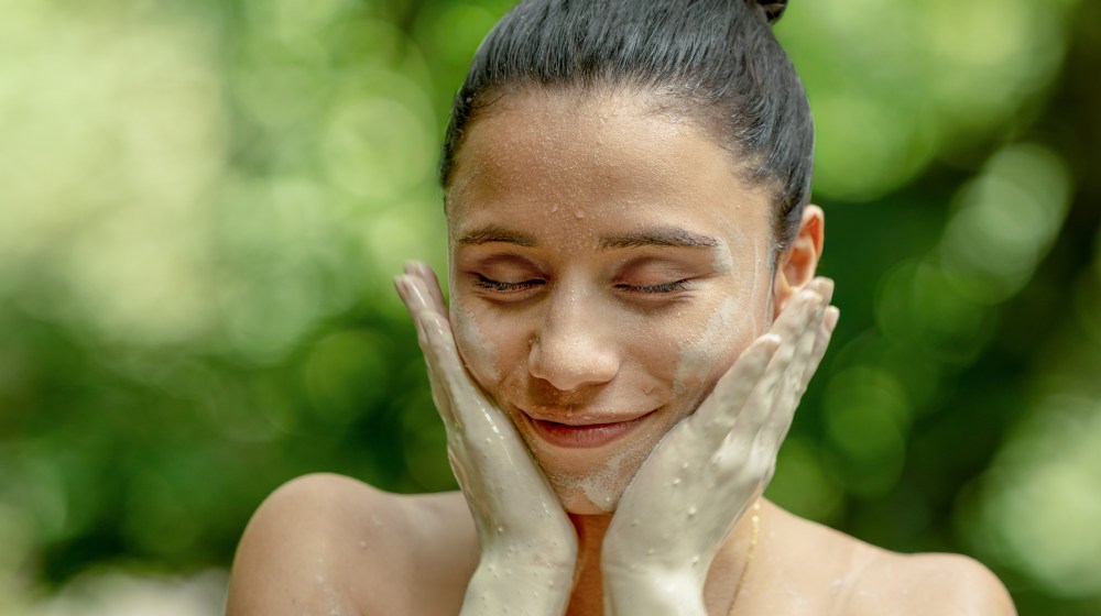 Person with clay mask on face, smiling with closed eyes, hands on cheeks, green background.