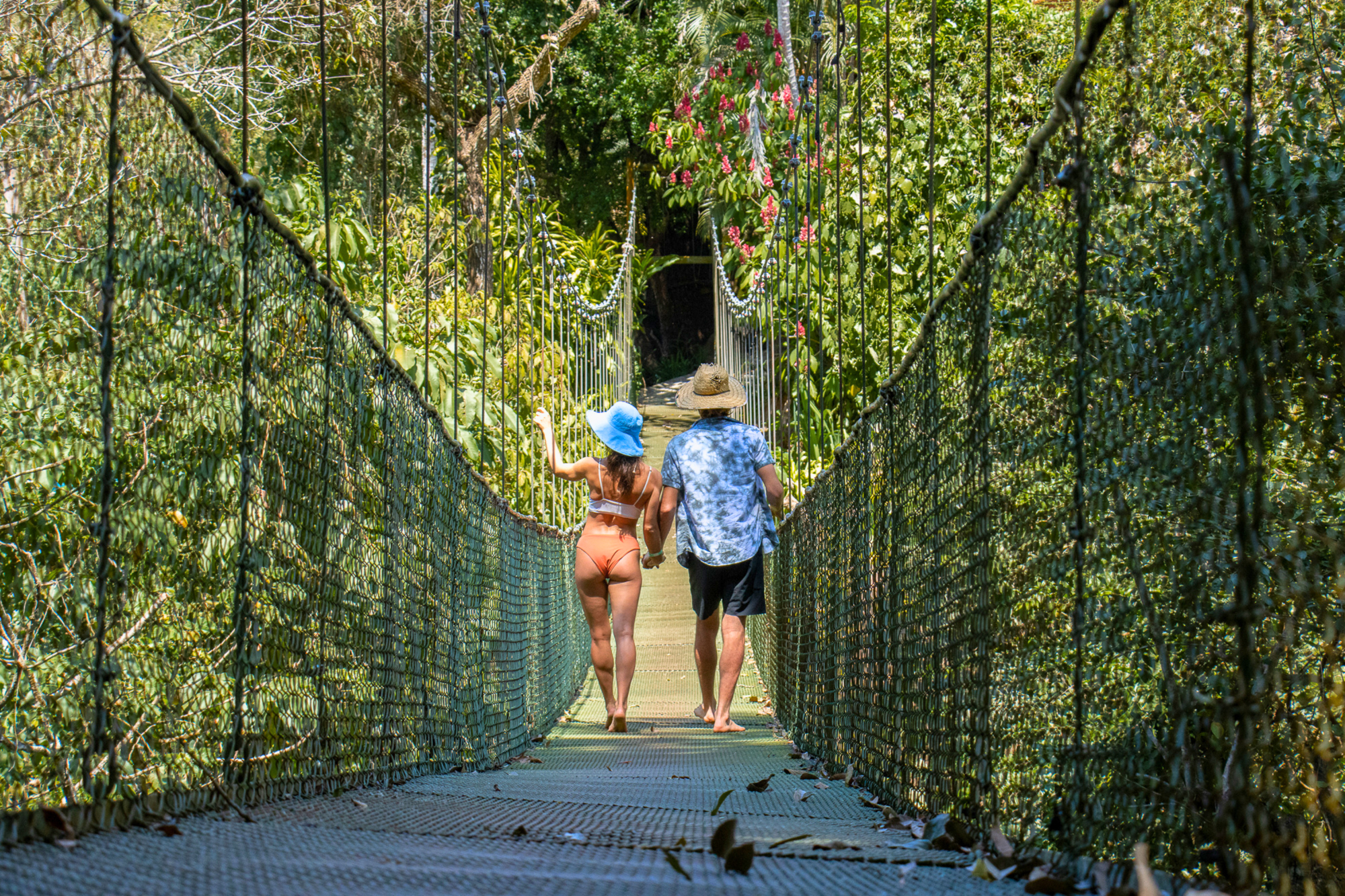 Dos personas con sombreros caminan por un puente colgante rodeado de exuberante vegetación.