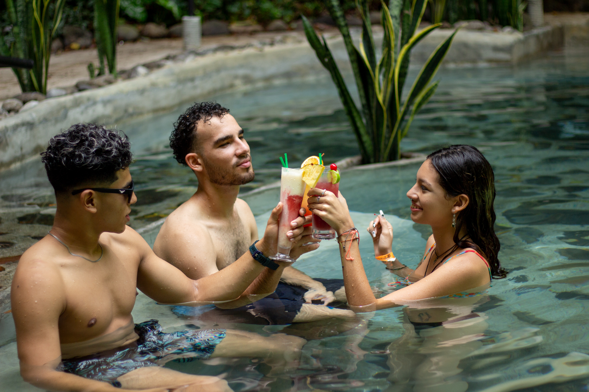 Tres personas en una piscina, sosteniendo bebidas coloridas y brindando.