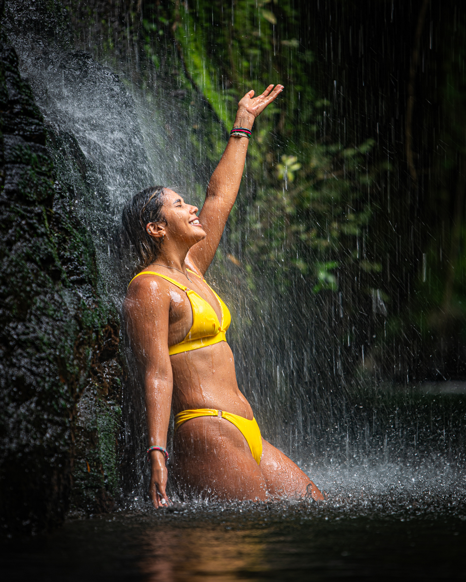 Mujer en bikini amarillo disfrutando de una cascada, estirando un brazo hacia arriba, rodeada de vegetación.