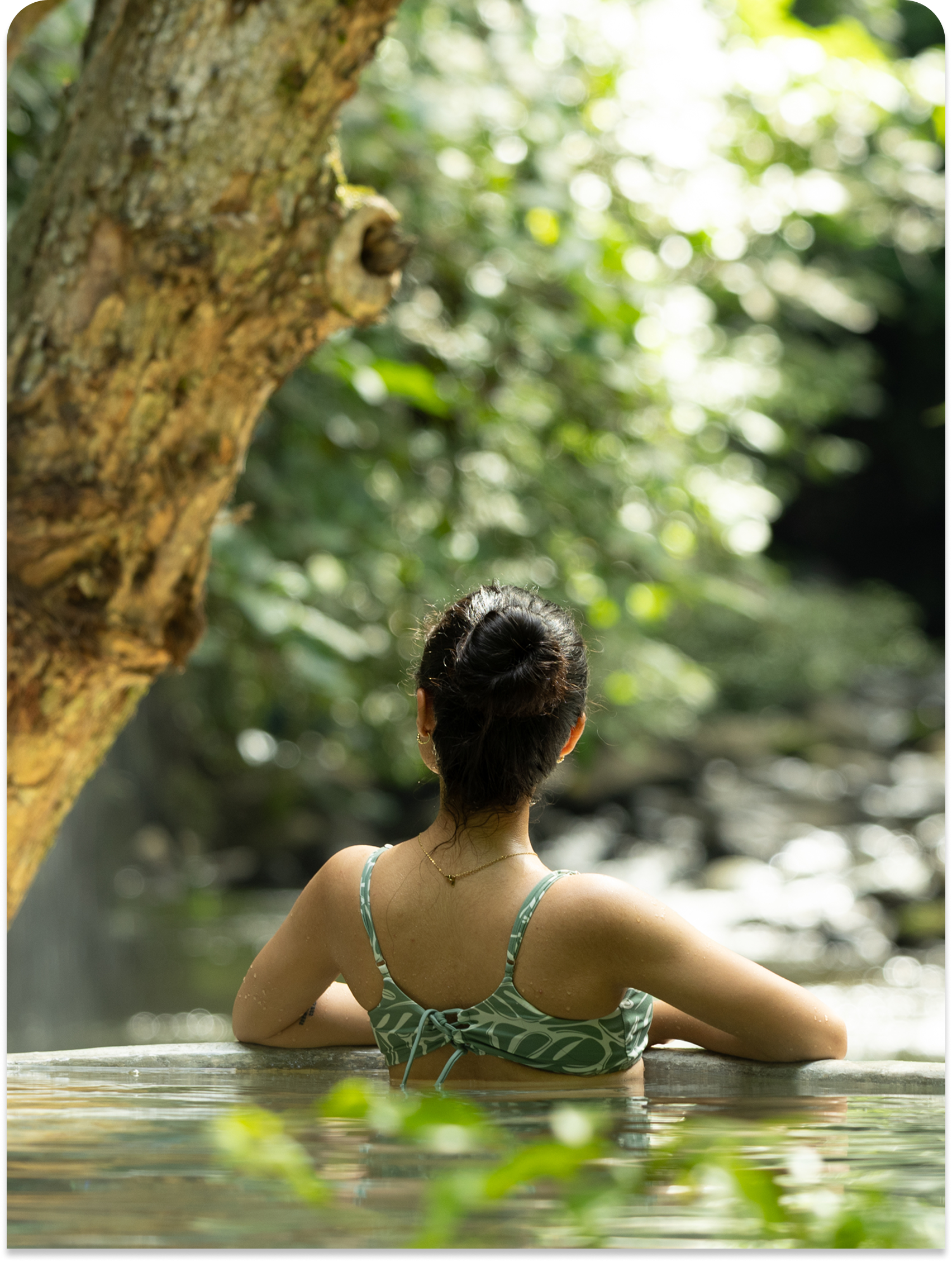 Mujer en bañador verde sentada en una piscina natural rodeada de exuberantes árboles.