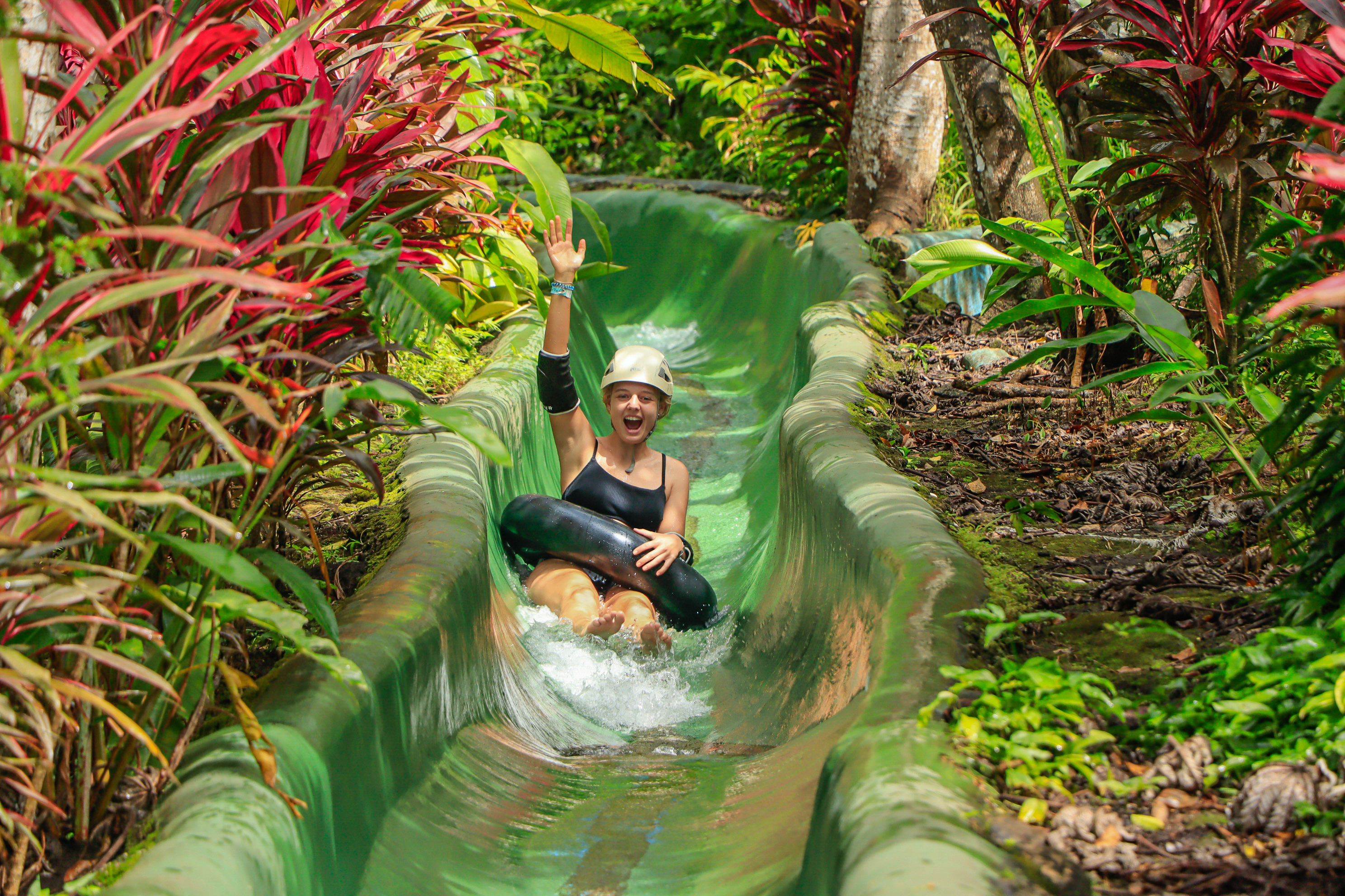 Persona en bañador negro deslizándose por un tobogán verde rodeado de plantas exuberantes.