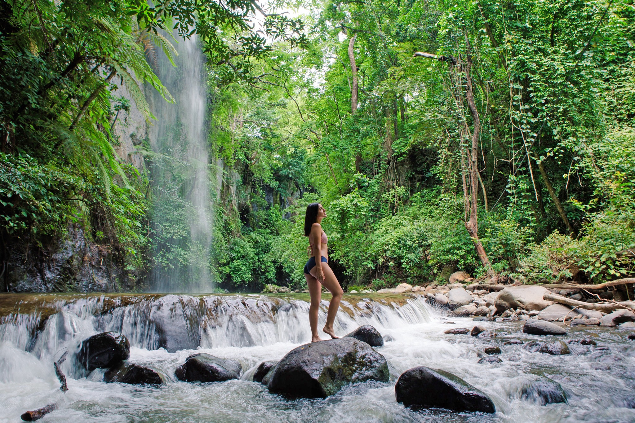 Person stands on rocks near a waterfall in a lush, green forest.
